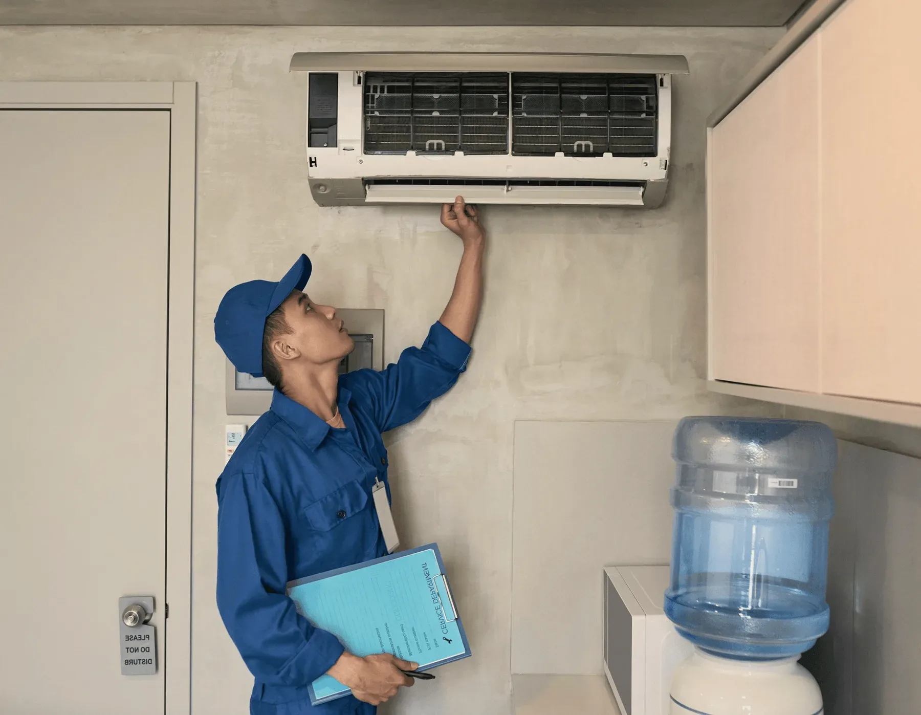 A technician in blue reaching up to a wall unit indoors.