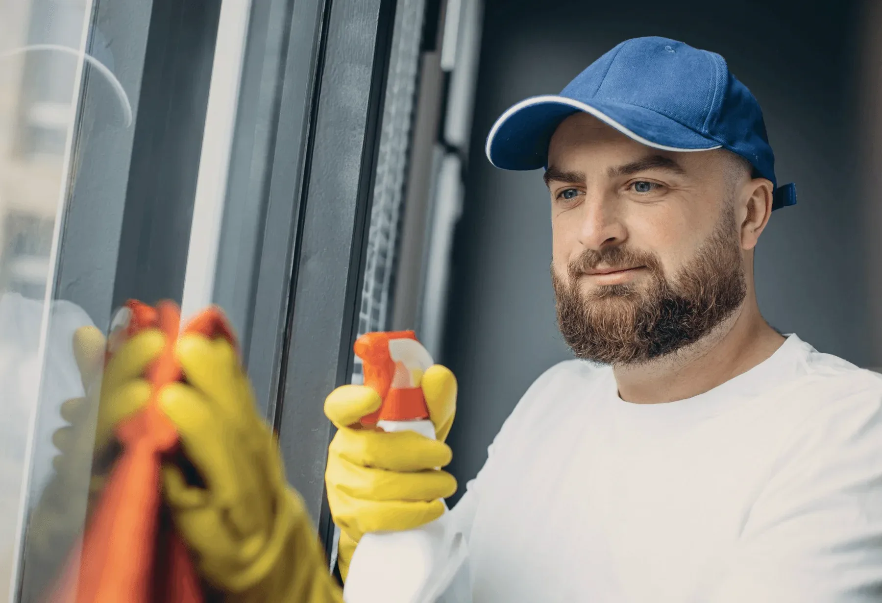 A bearded cleaner in a blue cap spraying a window.