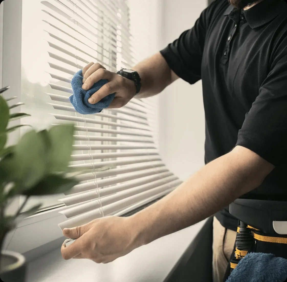 A cleaner in black wiping window blinds with a blue cloth.