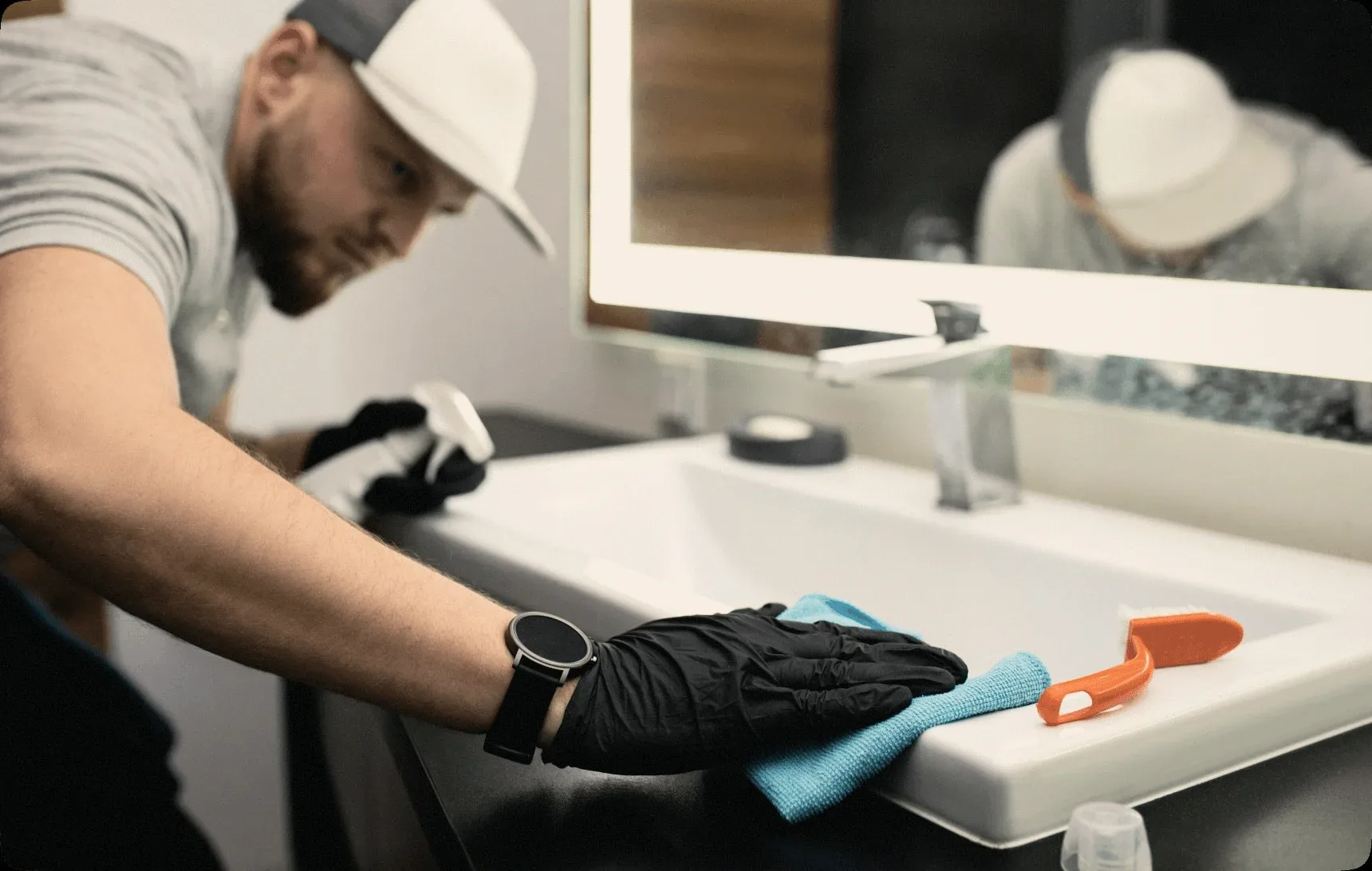A cleaner in a white cap wiping a bathroom sink.