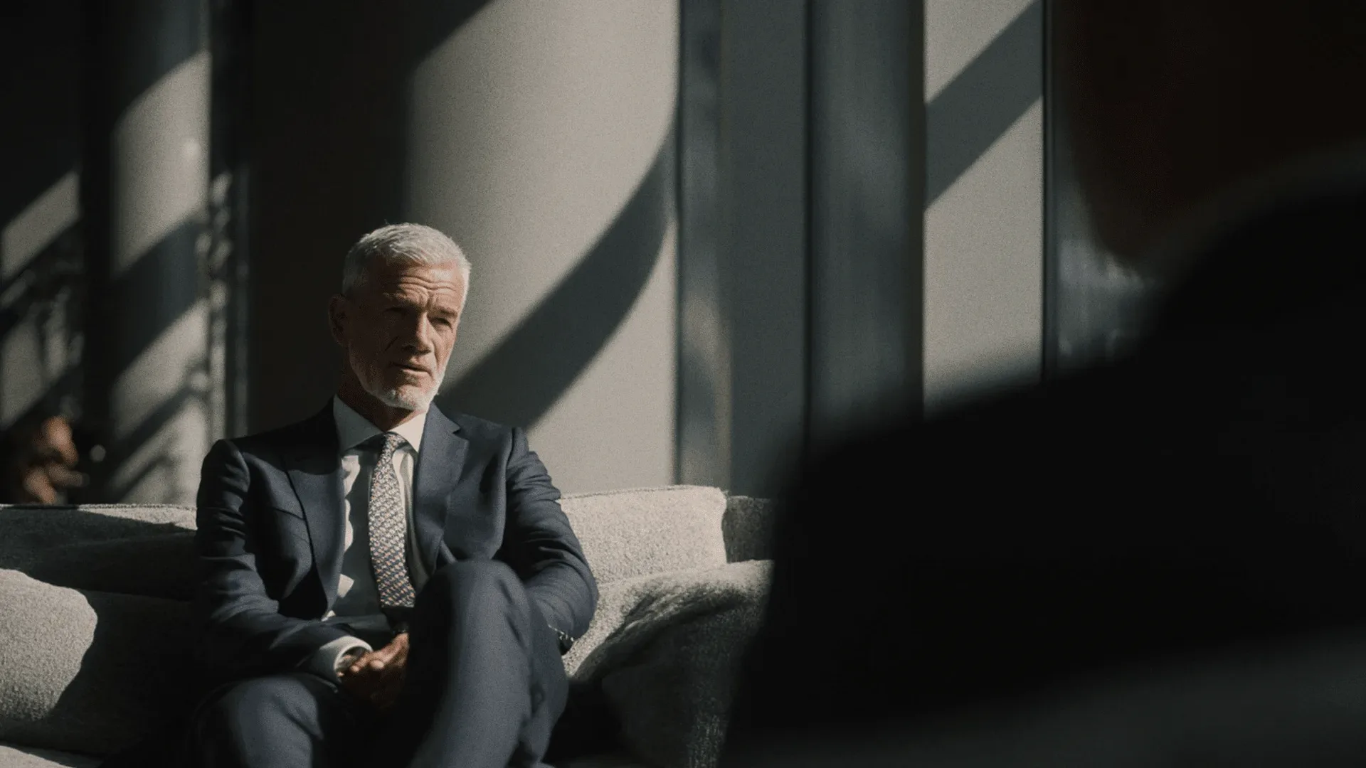 A man in a navy suit on a grey sofa in striped light.