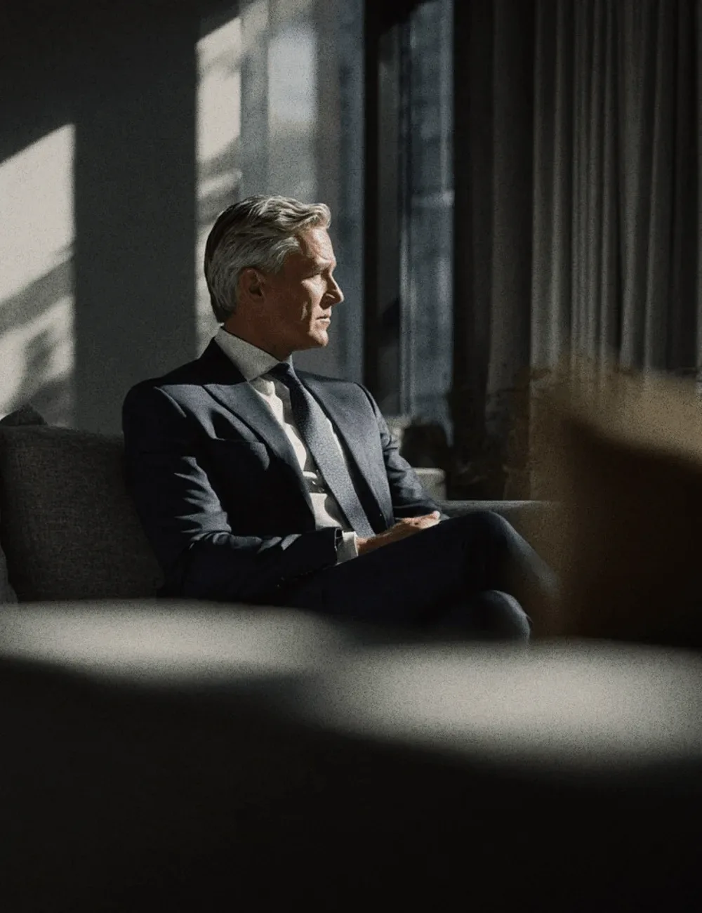 A man in a navy suit seated by a window.