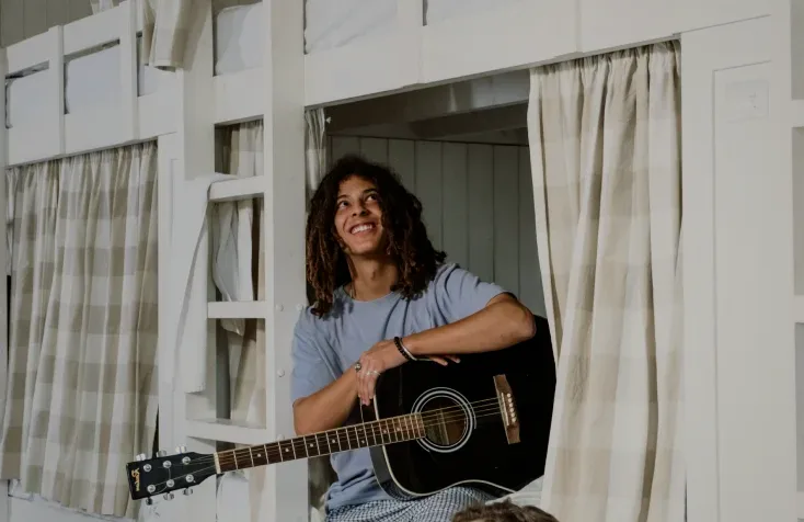 A man with a guitar smiling in a curtained bunk bed.