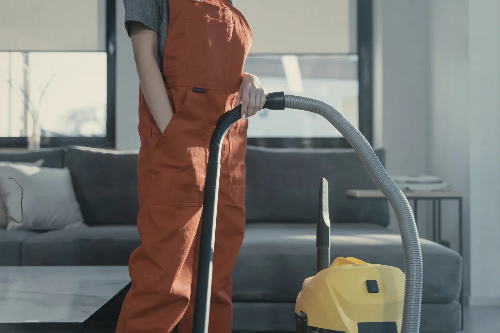 A cleaner in red overalls vacuuming beside a grey sofa.