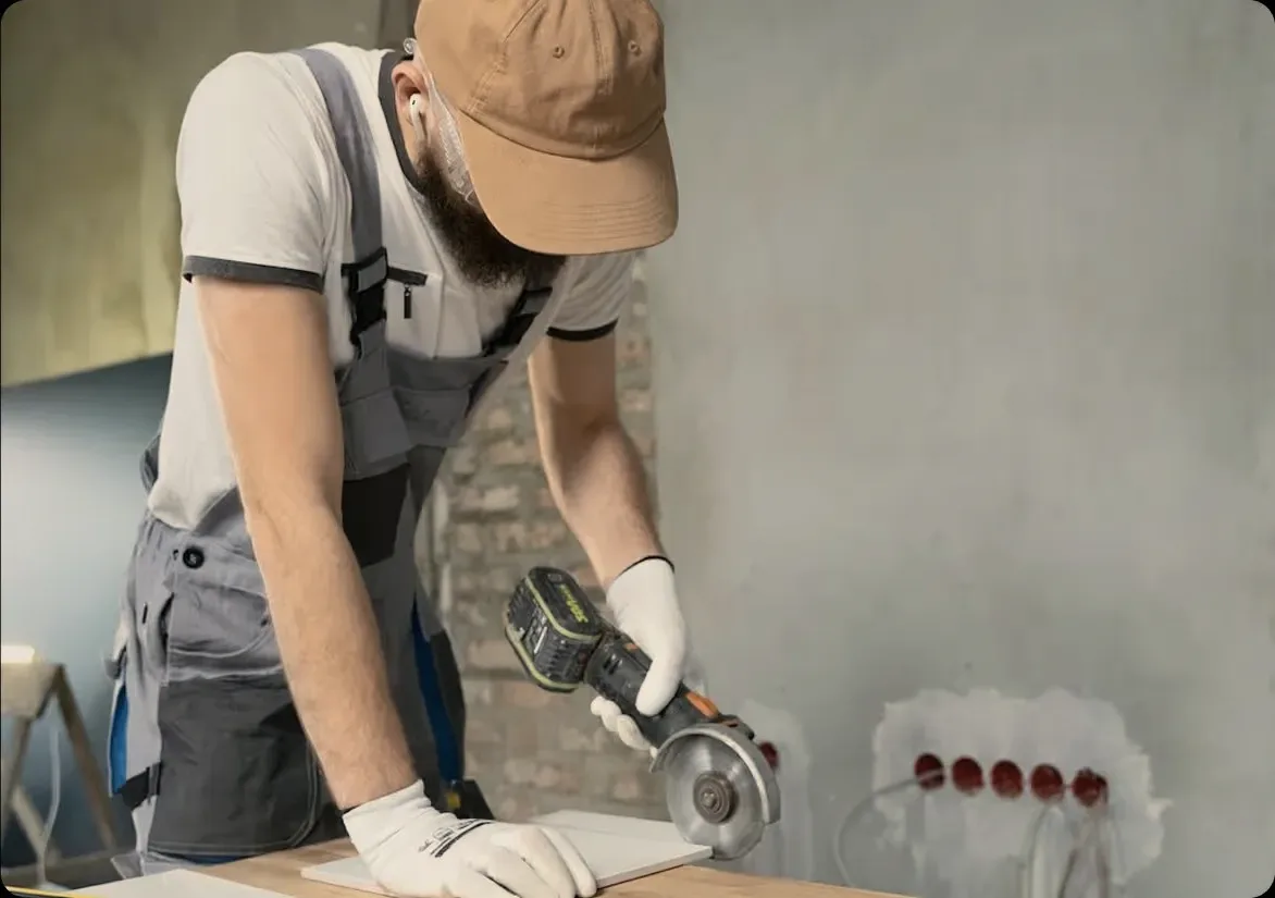 A worker in a cap and grey overalls cutting tile with a grinder.