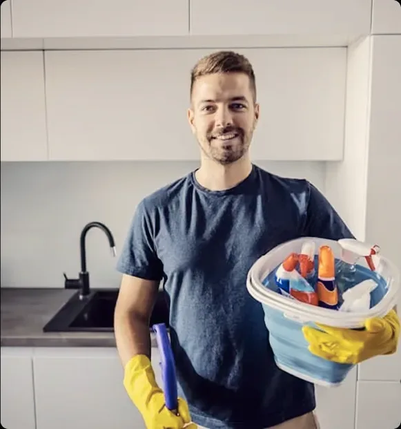 A smiling man in a navy shirt holding a basket of cleaning supplies.