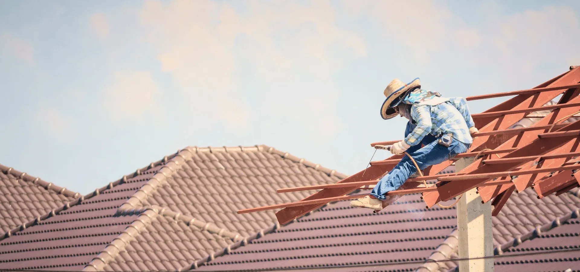 A roofer on red rafters under a soft sky.