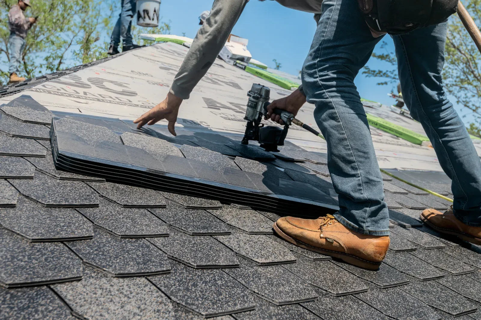Hands laying a new shingle on a roof deck, a worker's boots in frame beside it.