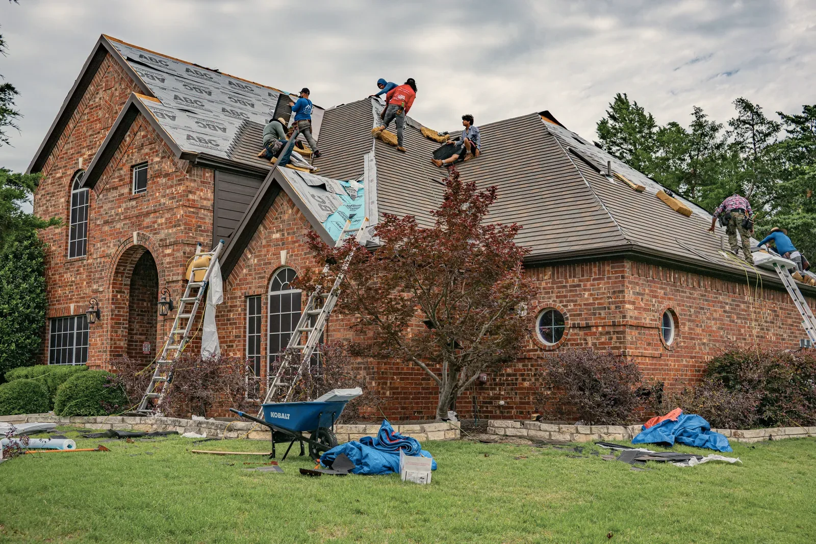 A crew re-roofing a brick house, ladders up and materials out on the lawn.
