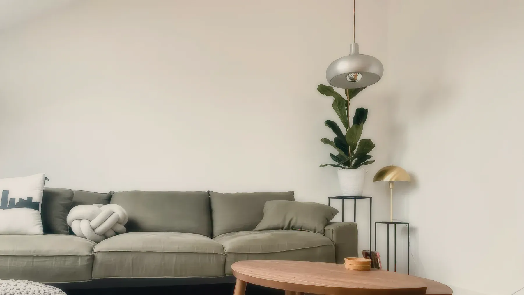 A calm living room: a green sofa, a round wooden table, a tall plant and a pendant lamp.