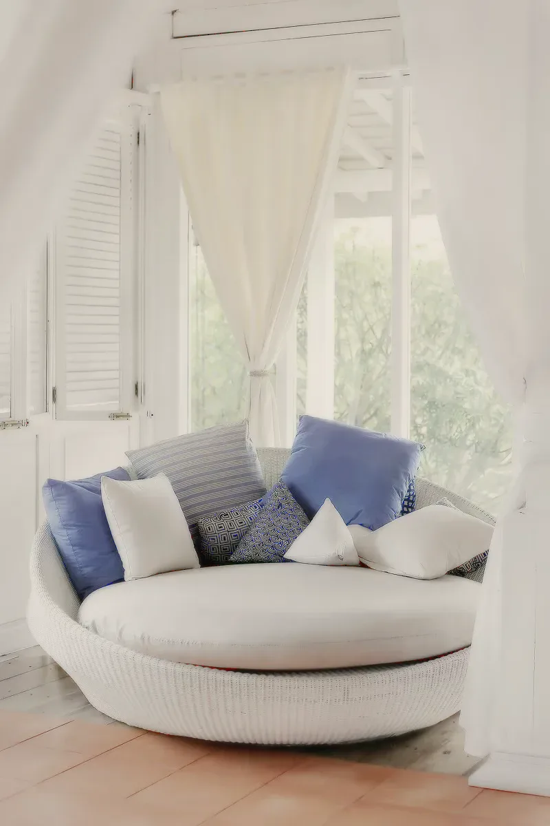 A round white wicker daybed with blue and white cushions by a curtained window.