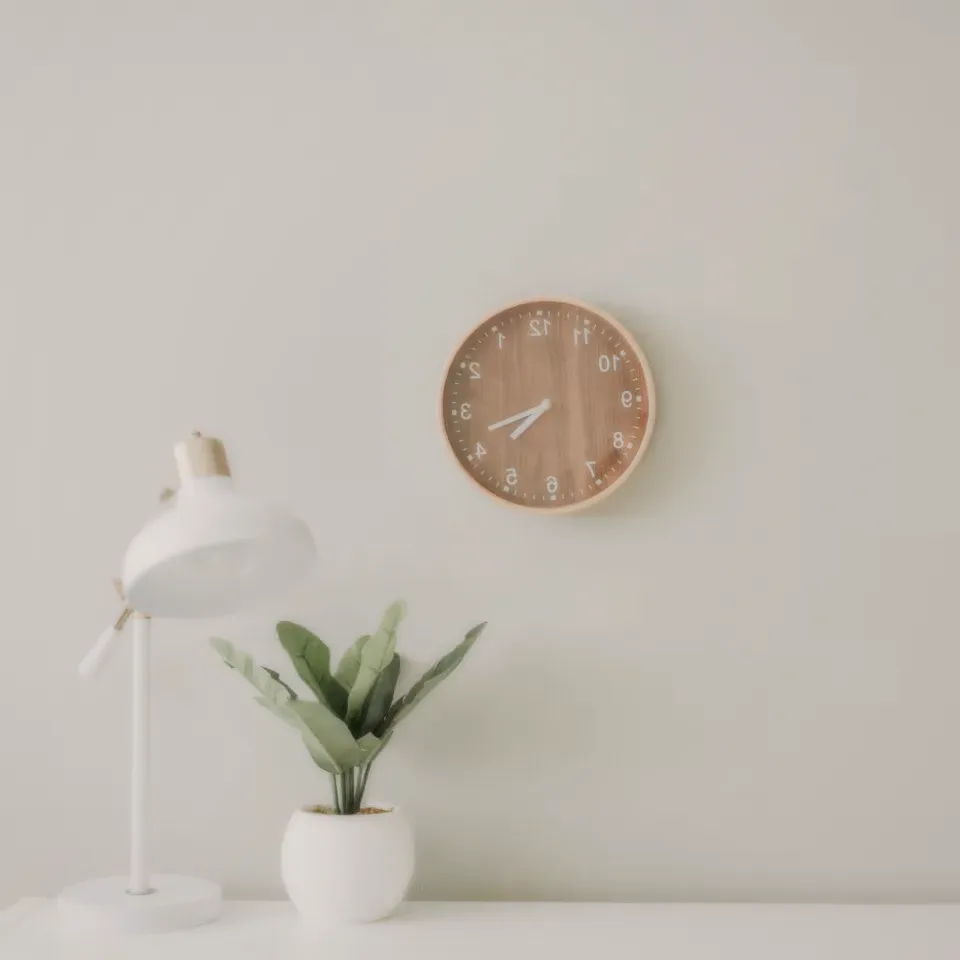 A round wooden clock over a white desk with a lamp and a plant.