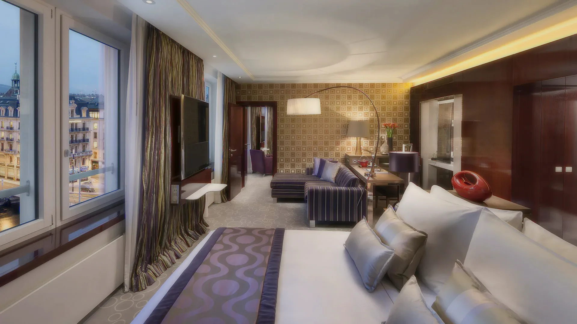 A hotel suite with a bed, a striped sofa, an arched lamp and a city view.