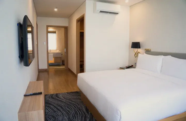 A white bedroom with a double bed, a wall-mounted screen and a small hallway.