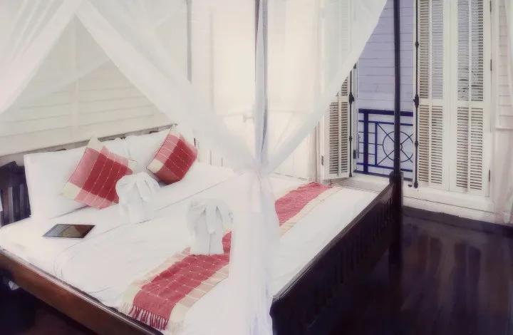 A four-poster bed with white netting and red cushions by a balcony door.